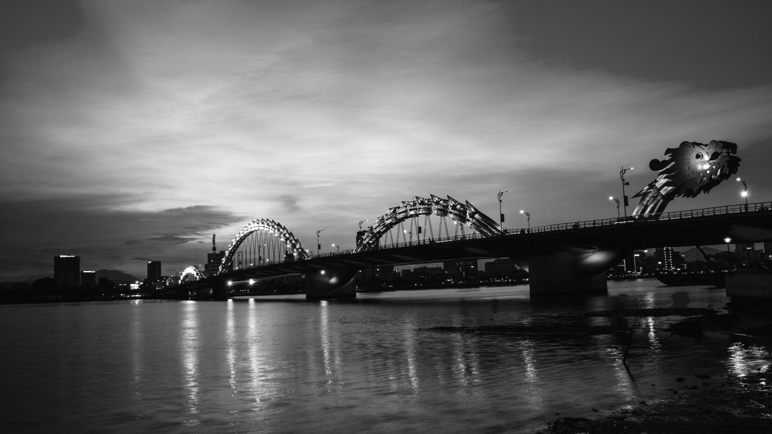 Dragon-shaped bridge over calm water at dusk with a city skyline in the background under a cloudy sky.