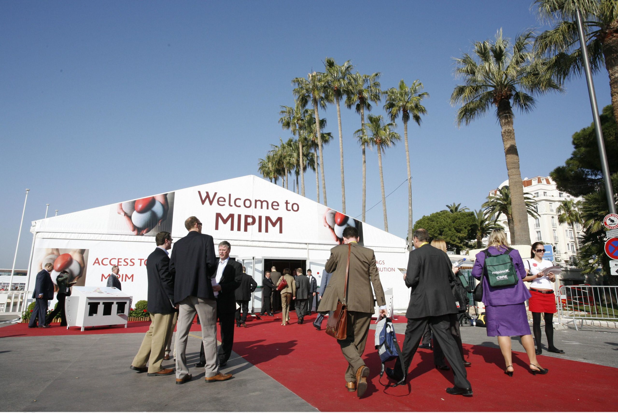Business professionals entering a large tent at an event with palm trees and a welcome sign in the background.