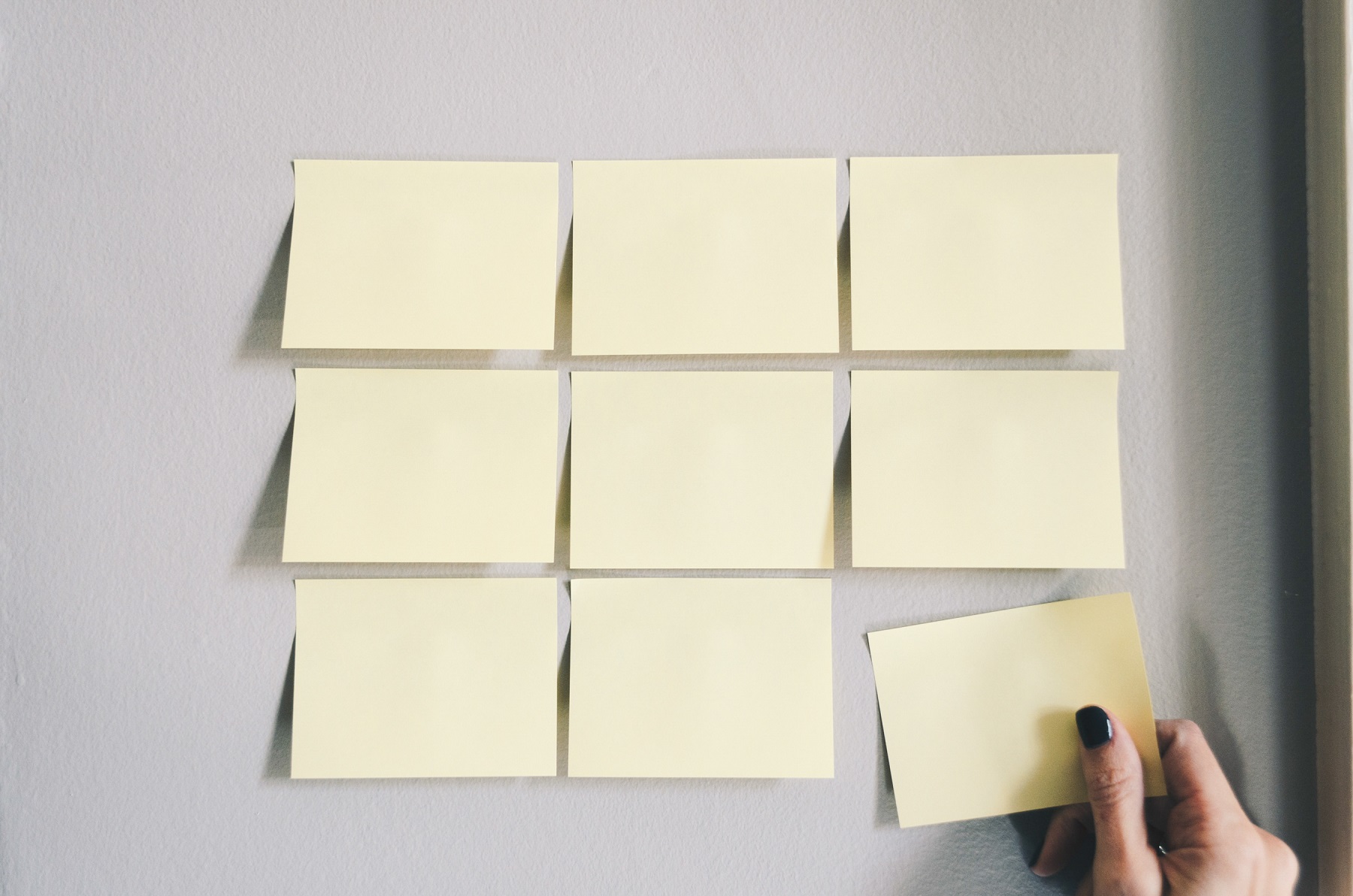 Hand placing a yellow sticky note on a wall with several other blank sticky notes arranged in a grid pattern.