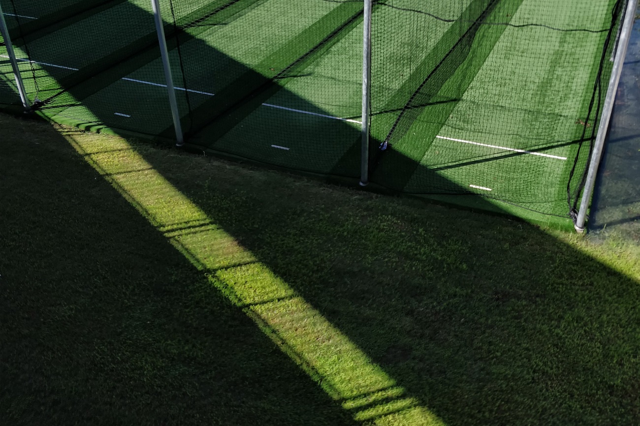 Indoor sports facility with green artificial turf and netting, creating shadows on the grass surface.
