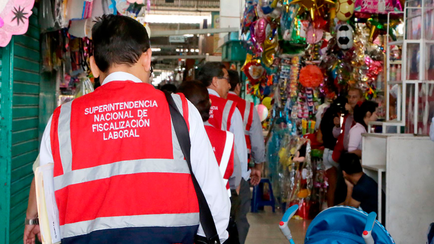 Inspectors wearing red vests walking through a marketplace with various colorful items on display.