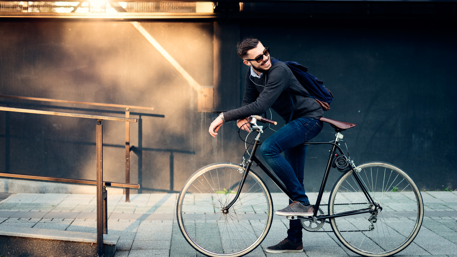 Man in casual attire riding a bicycle along a sidewalk with a railing in the background.