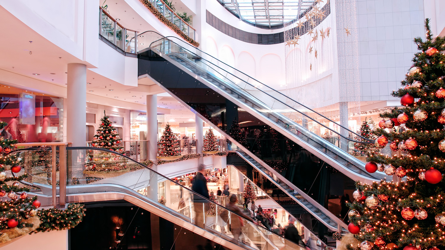 Shopping mall interior with escalators and decorated Christmas trees. Shoppers visible in the background.