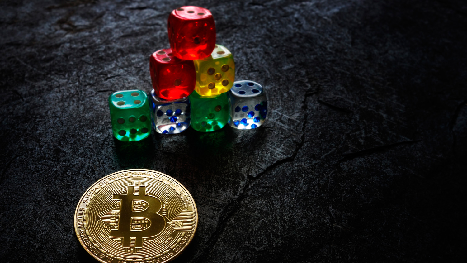 Bitcoin coin positioned in front of stacked colored dice on a textured dark surface.