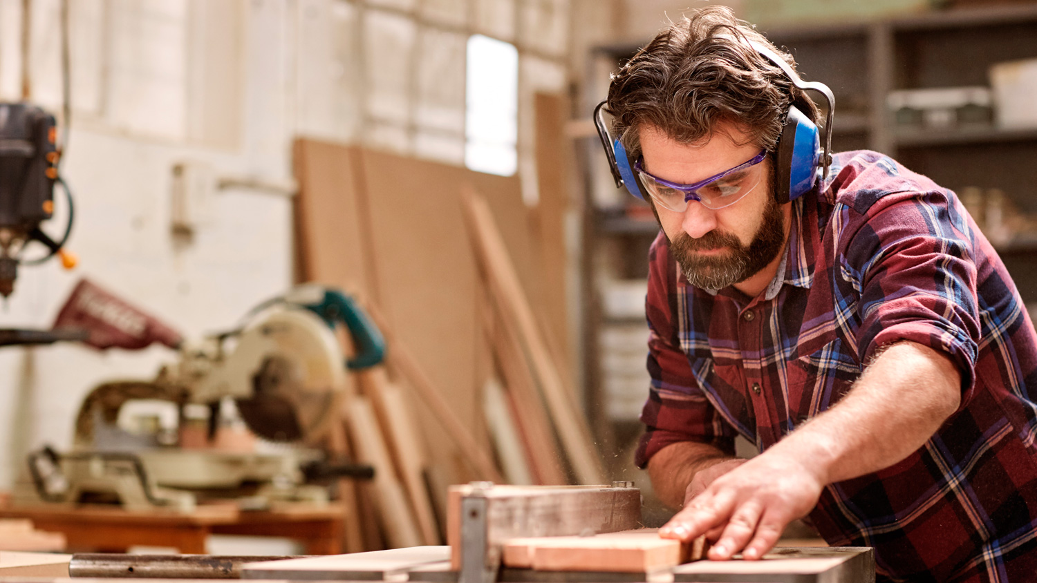 Craftsman using power tools to shape wood in a well-lit workshop, wearing safety goggles and hearing protection.