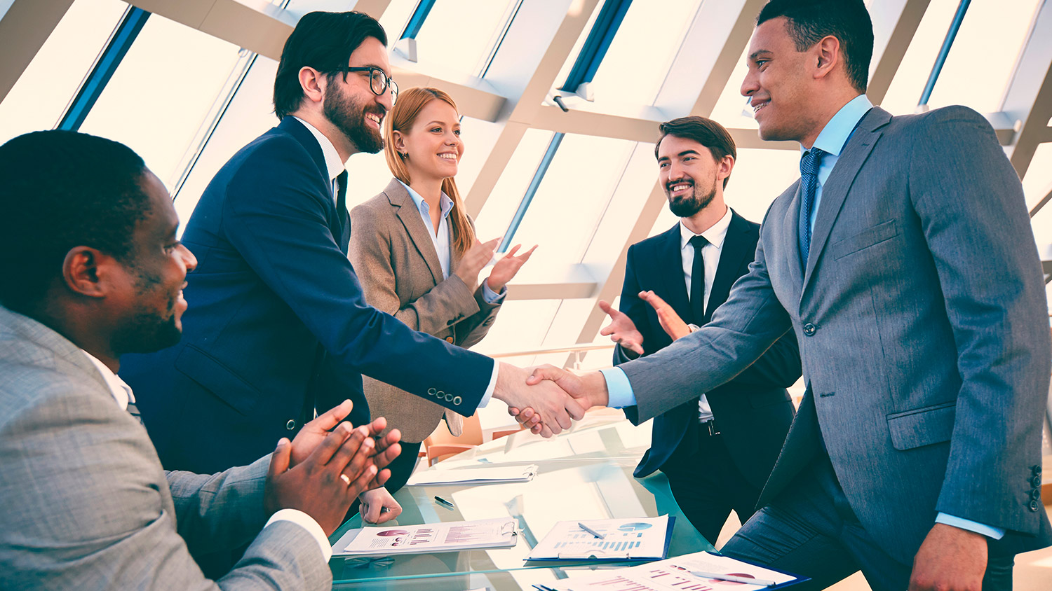 Two attorneys shaking hands in a bright conference room while colleagues applaud and review legal documents.