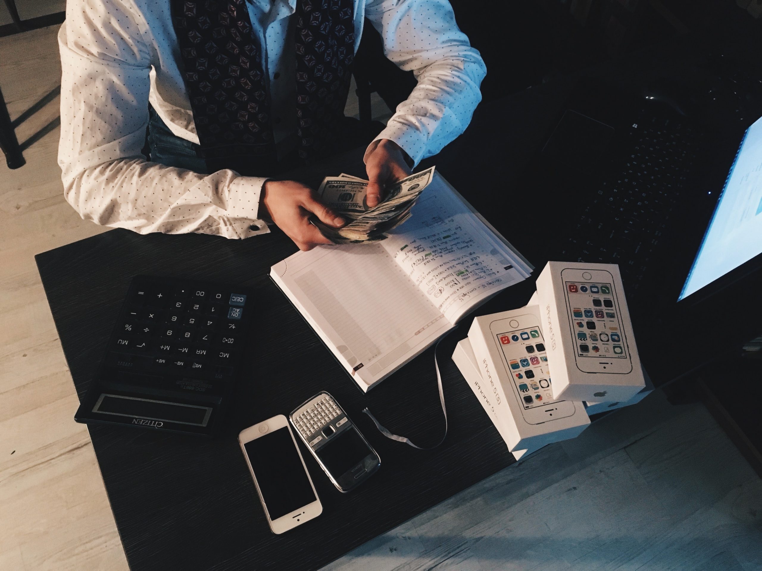 Person counting cash at a desk with a calculator, notepad, and mobile phones in a dimly lit office setting.
