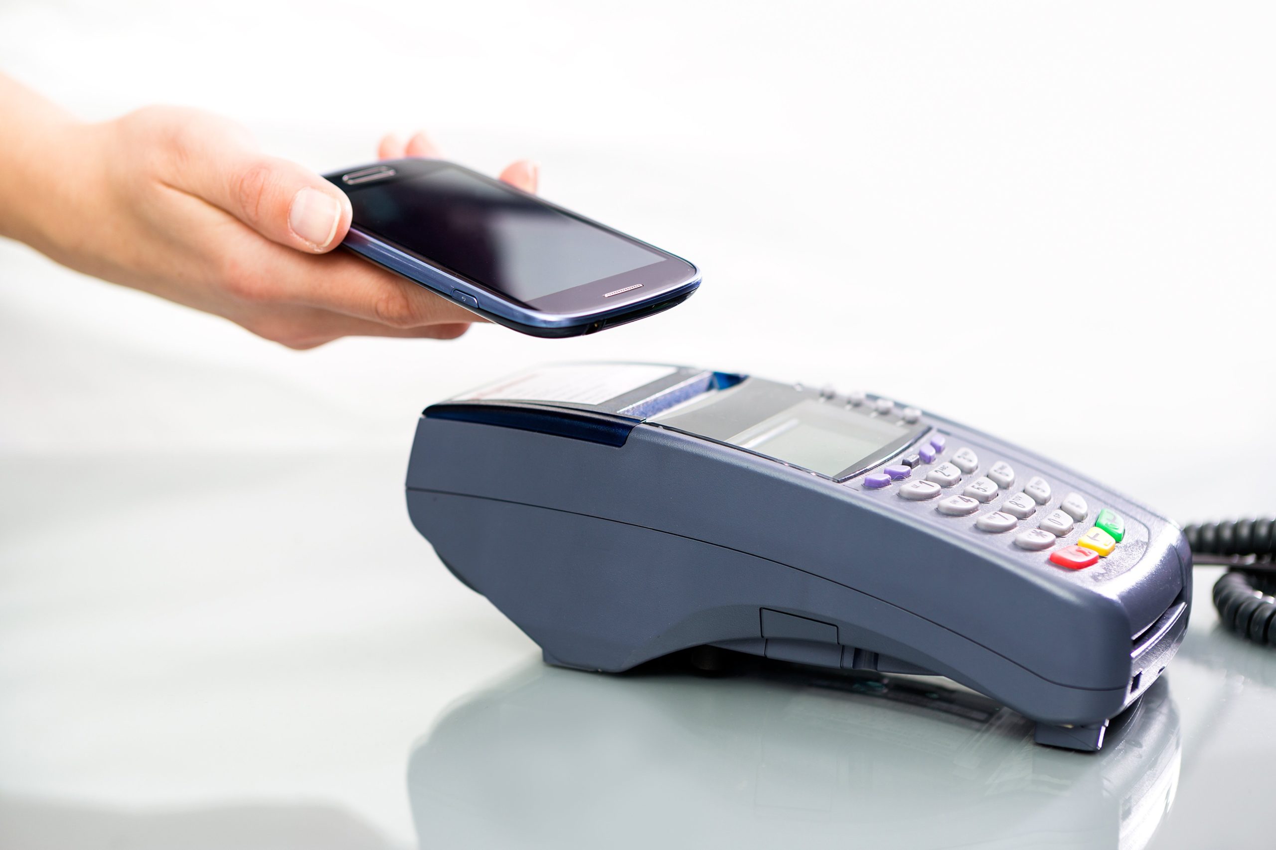 Hand holding a smartphone above a contactless payment terminal on a table.