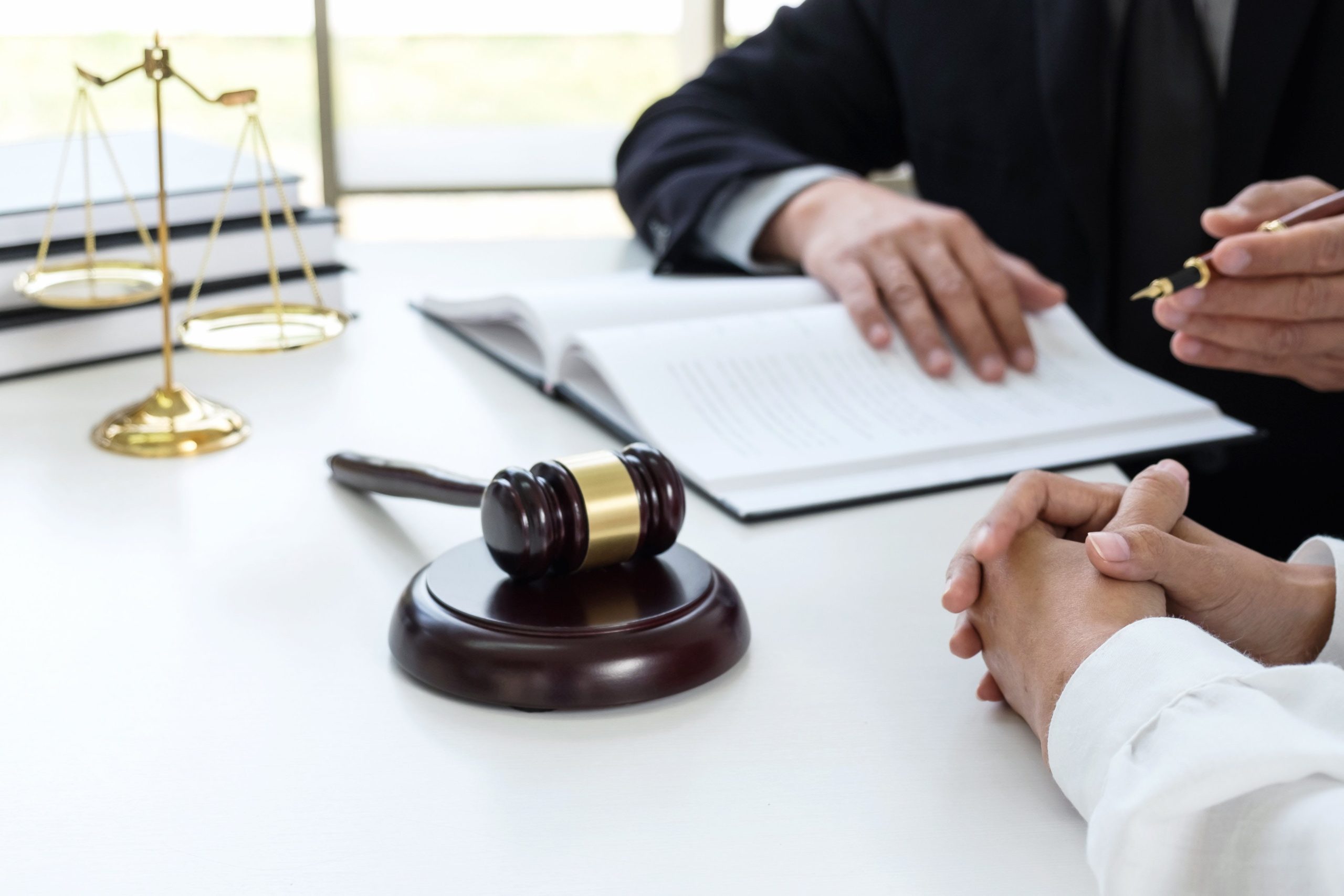 Attorney consulting with client at law firm desk, gavel and scales of justice visible, reviewing legal documents.