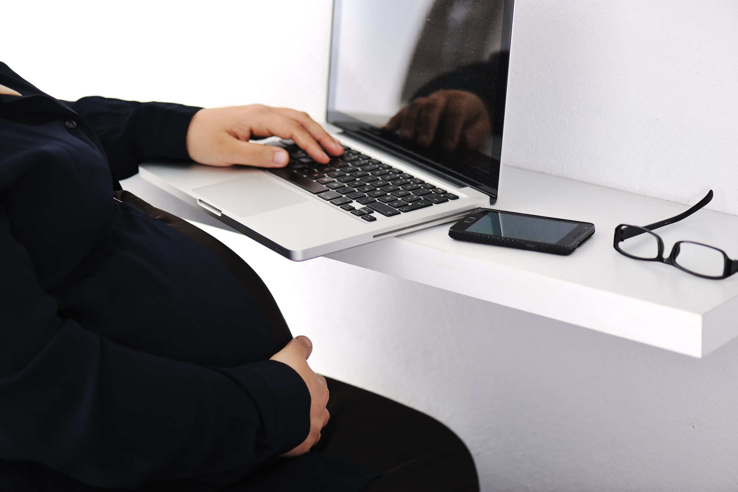 Pregnant individual typing on a laptop at a desk with a smartphone and glasses nearby.