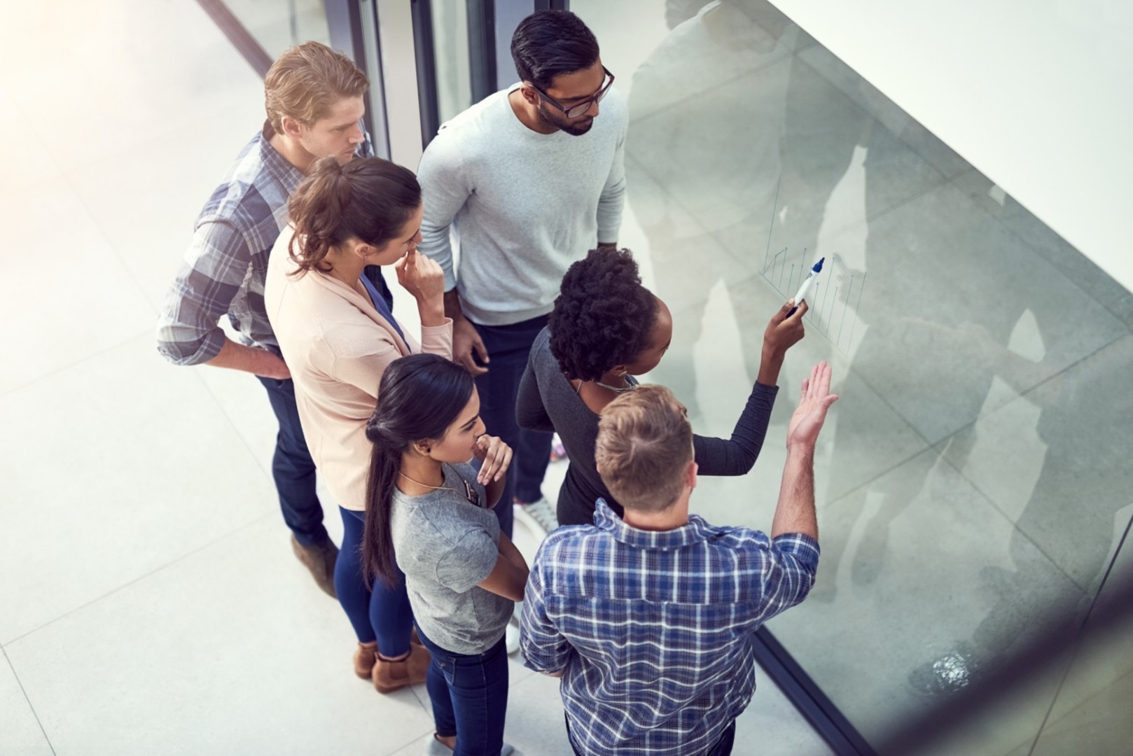 Group of six professionals collaborating at a glass wall, one using a marker to illustrate ideas.