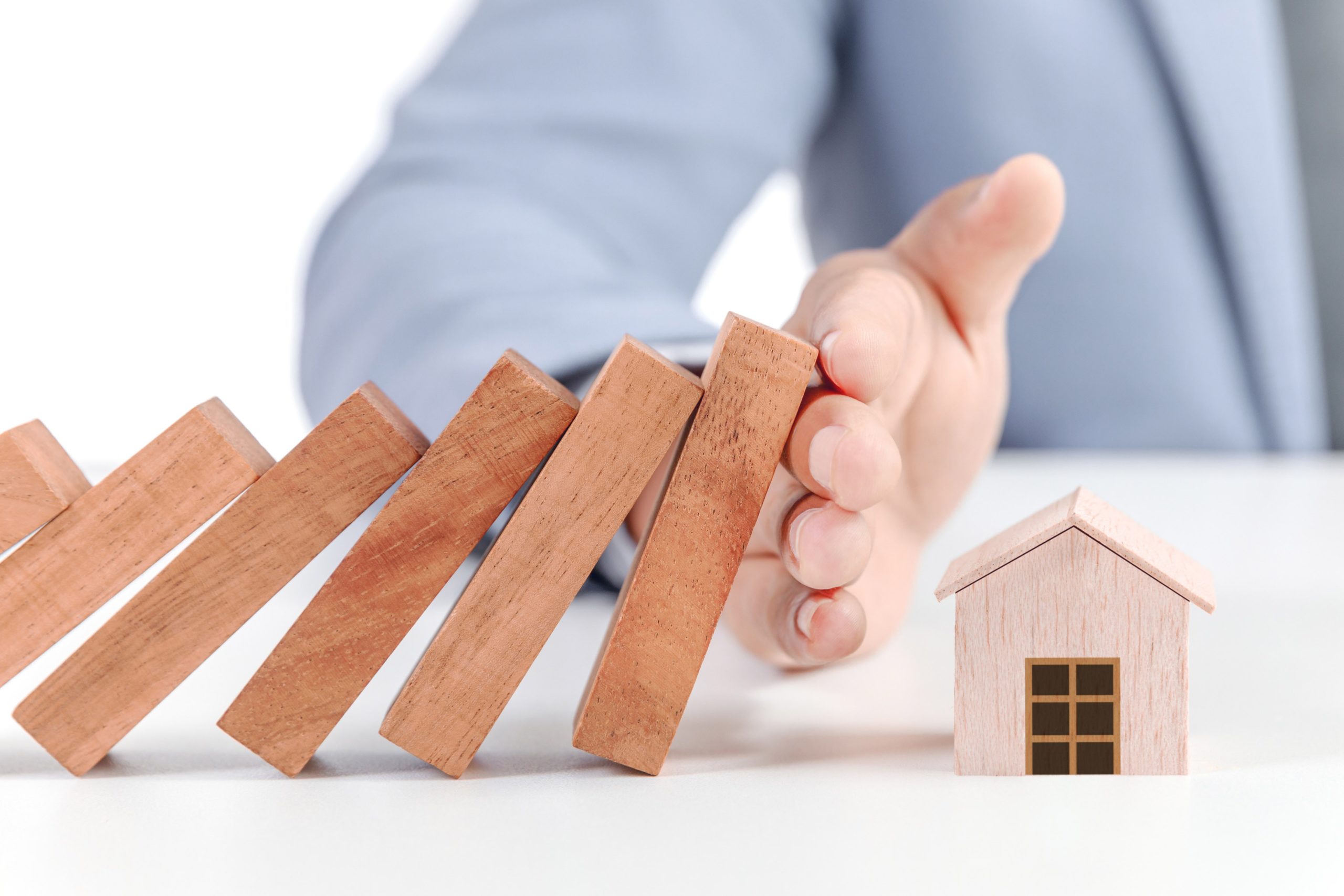 Hand positioned to prevent falling wooden blocks near a small house model, symbolizing risk management in real estate.