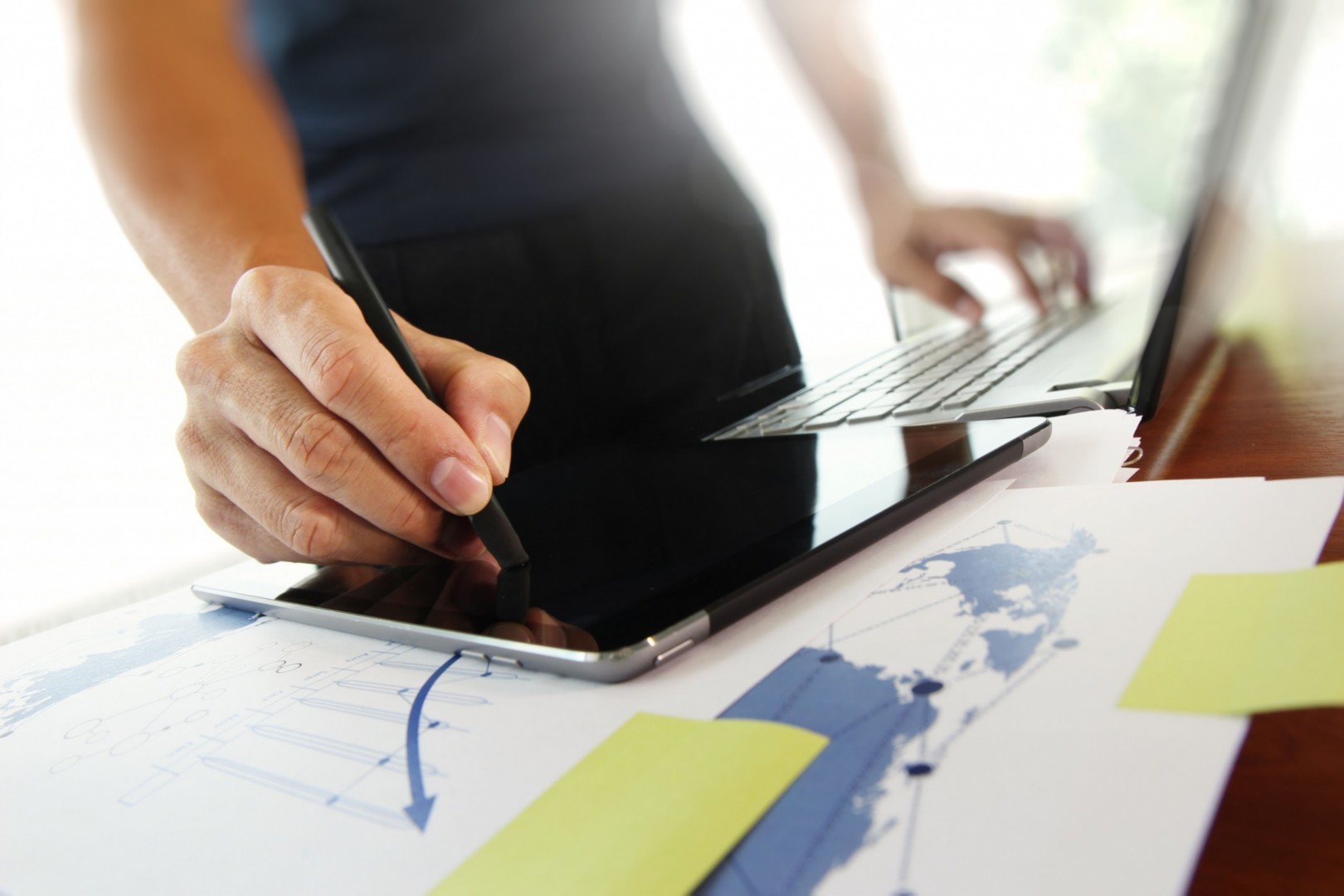 Professional using a stylus on a tablet while reviewing documents on a laptop at a desk with sticky notes.