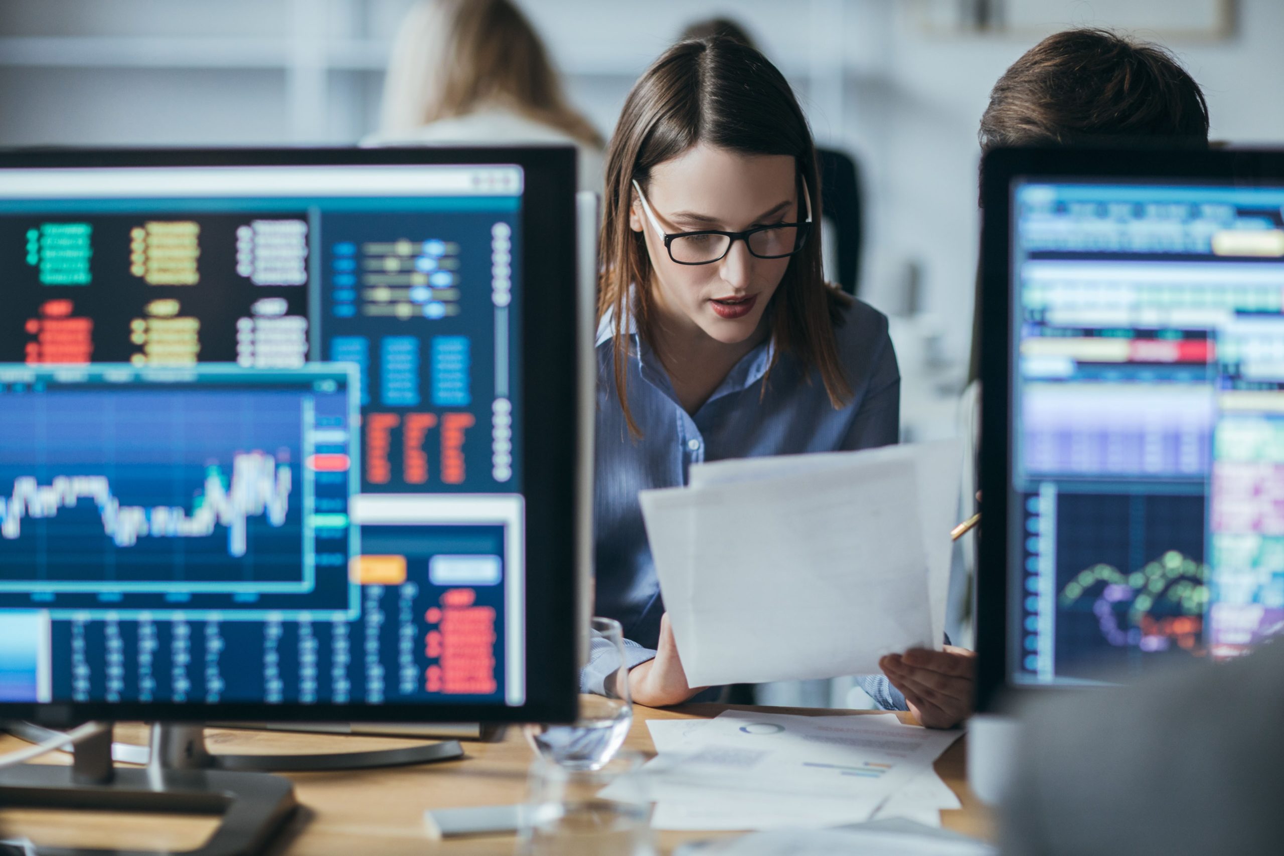 Business professional analyzing documents while reviewing data on multiple computer screens.