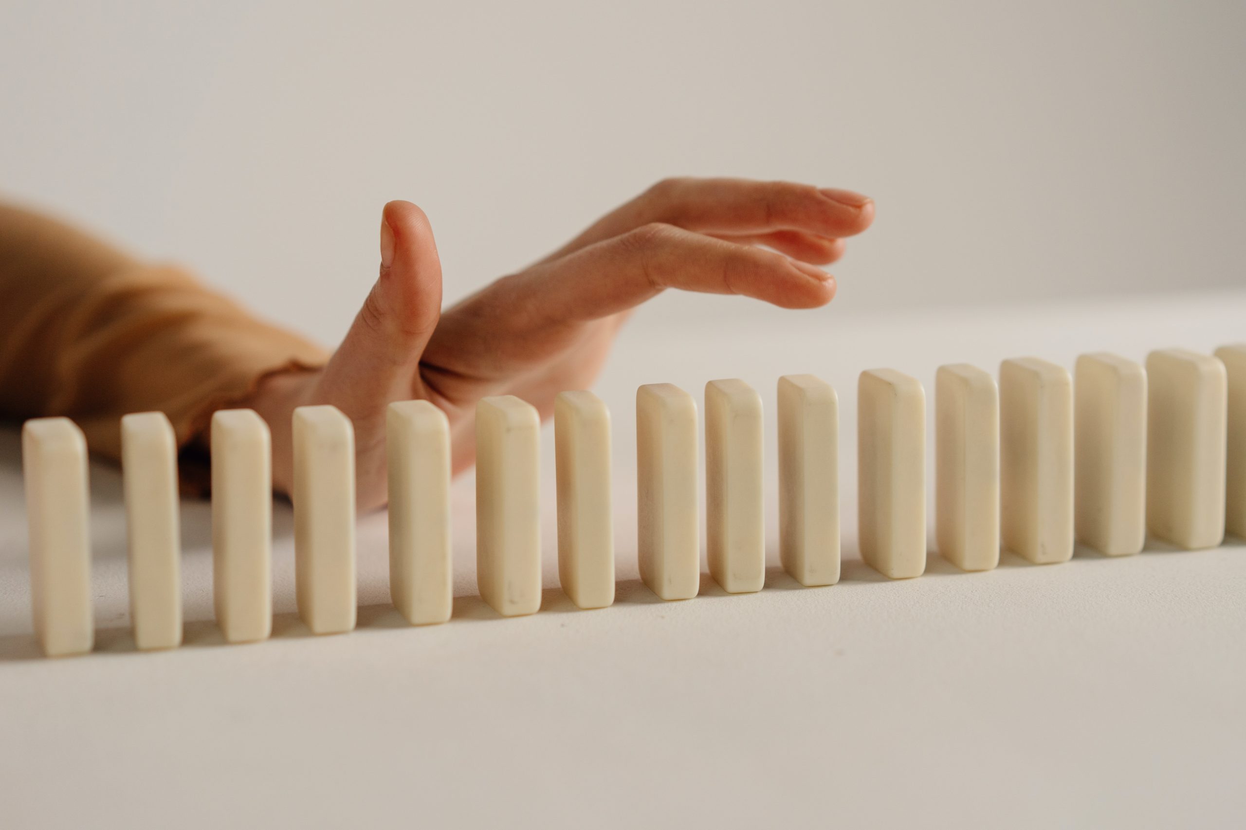 Hand reaching to topple a line of white dominoes on a flat surface.