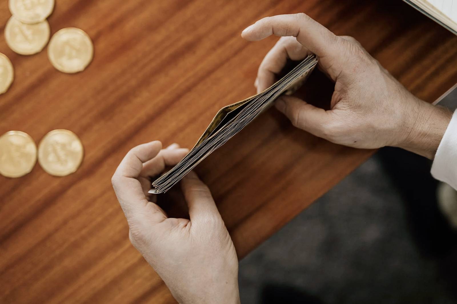 Person holding a stack of cash on a wooden table with coins nearby.