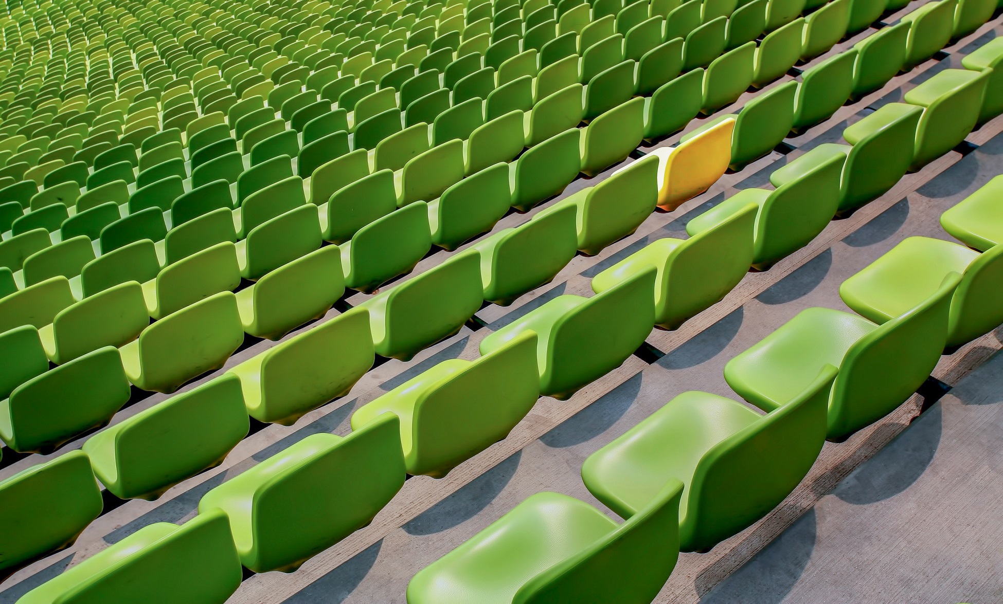 Empty green stadium seats arranged in rows, with one yellow seat prominently visible among the green ones.