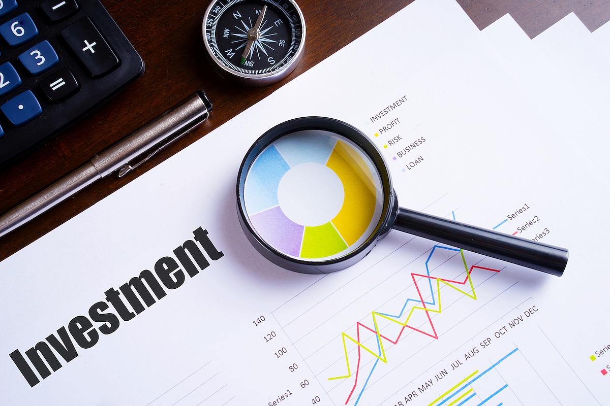 Magnifying glass on investment report with graphs, calculator, pen, and compass on wooden desk.