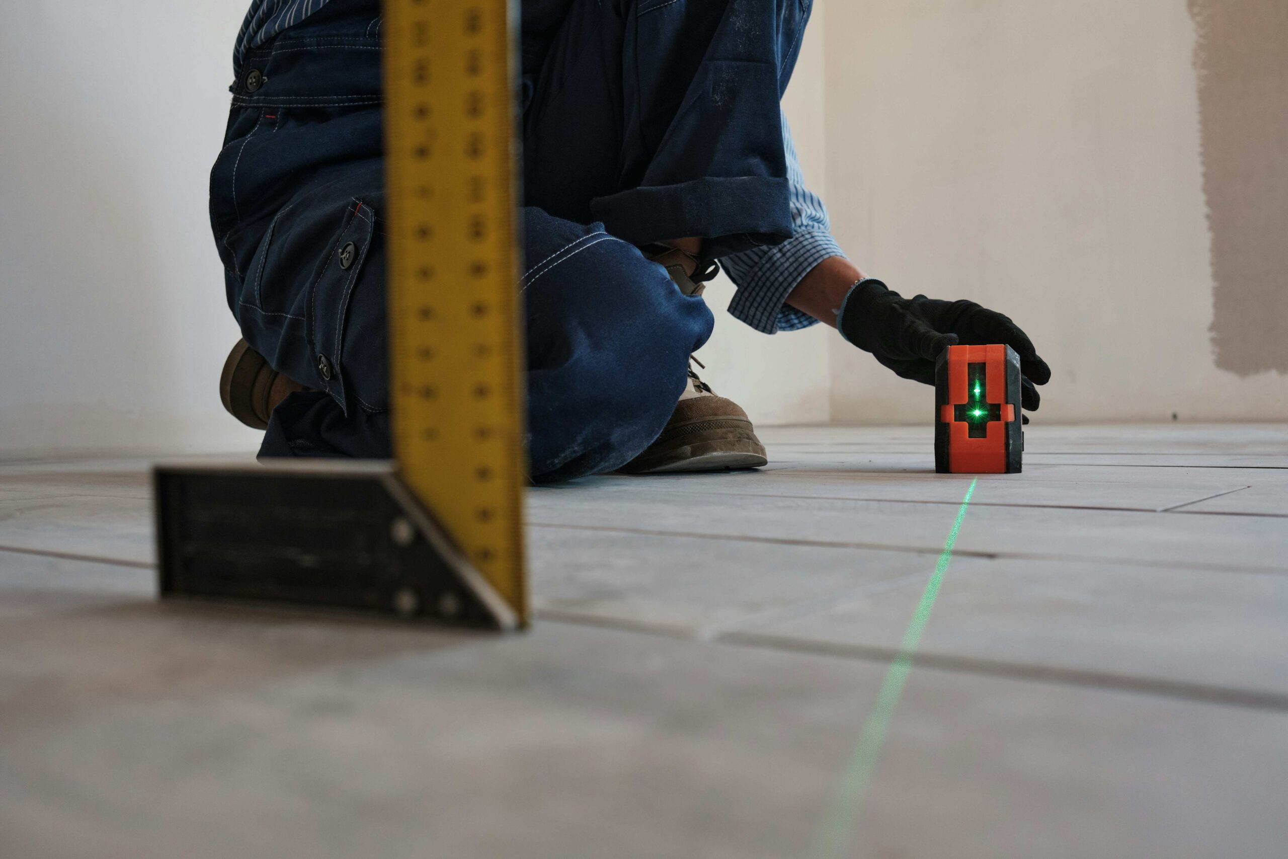 Worker measuring flooring with laser level tool and square in a partially finished room.