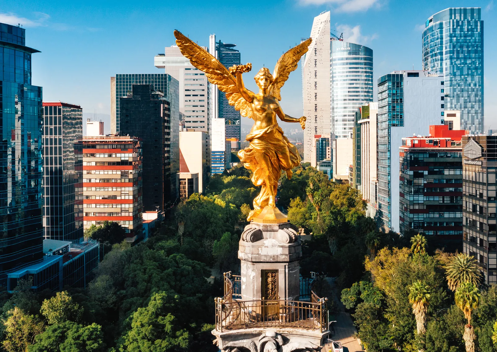 Golden statue of a winged figure atop a monument surrounded by urban buildings and greenery in a cityscape.