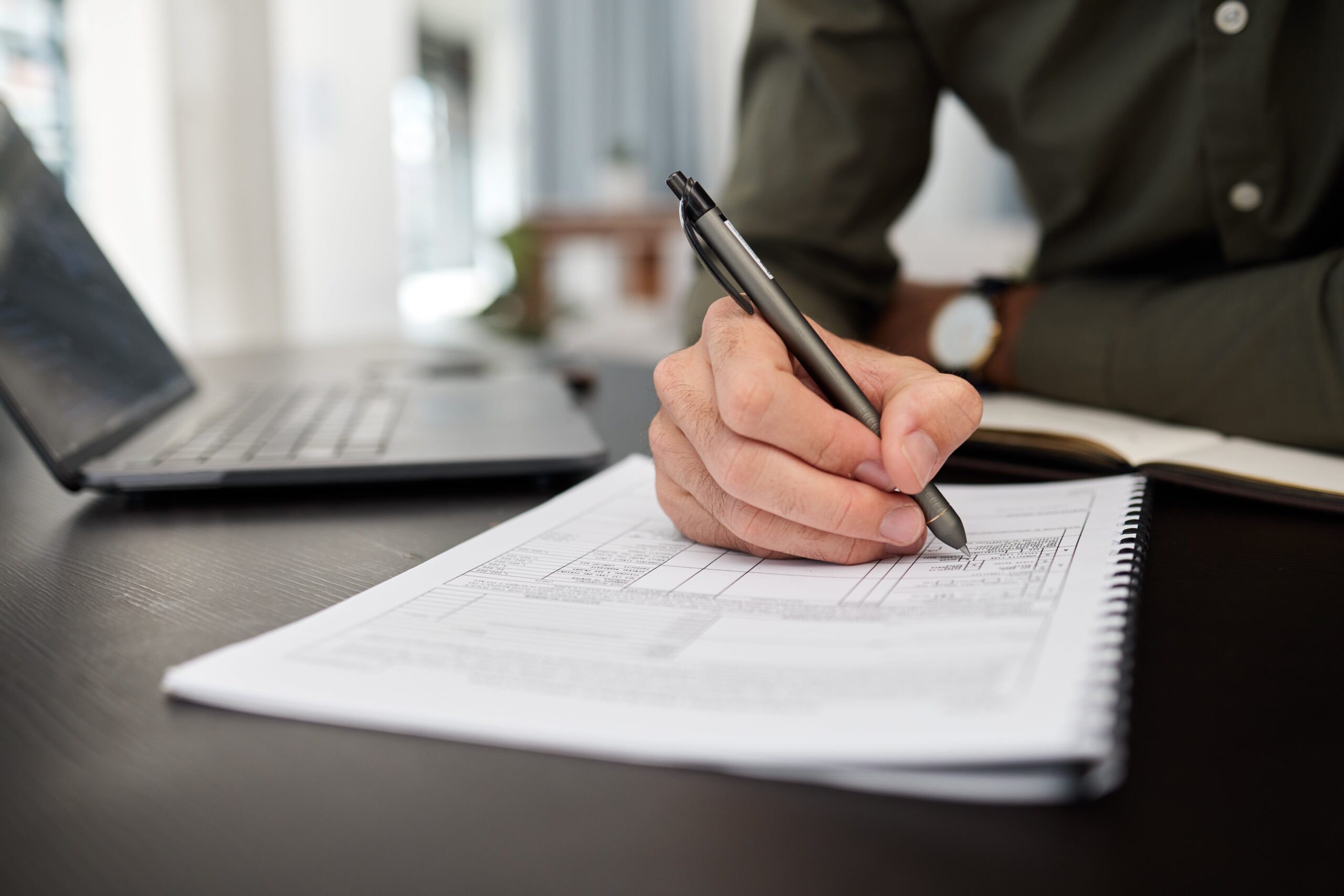 Attorney writing notes on legal documents with a laptop and notebook in an office setting.