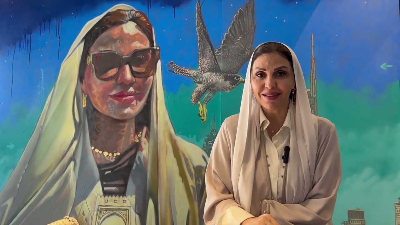 Woman in light-colored attire standing in front of a mural featuring a portrait and falcon in a vibrant setting.