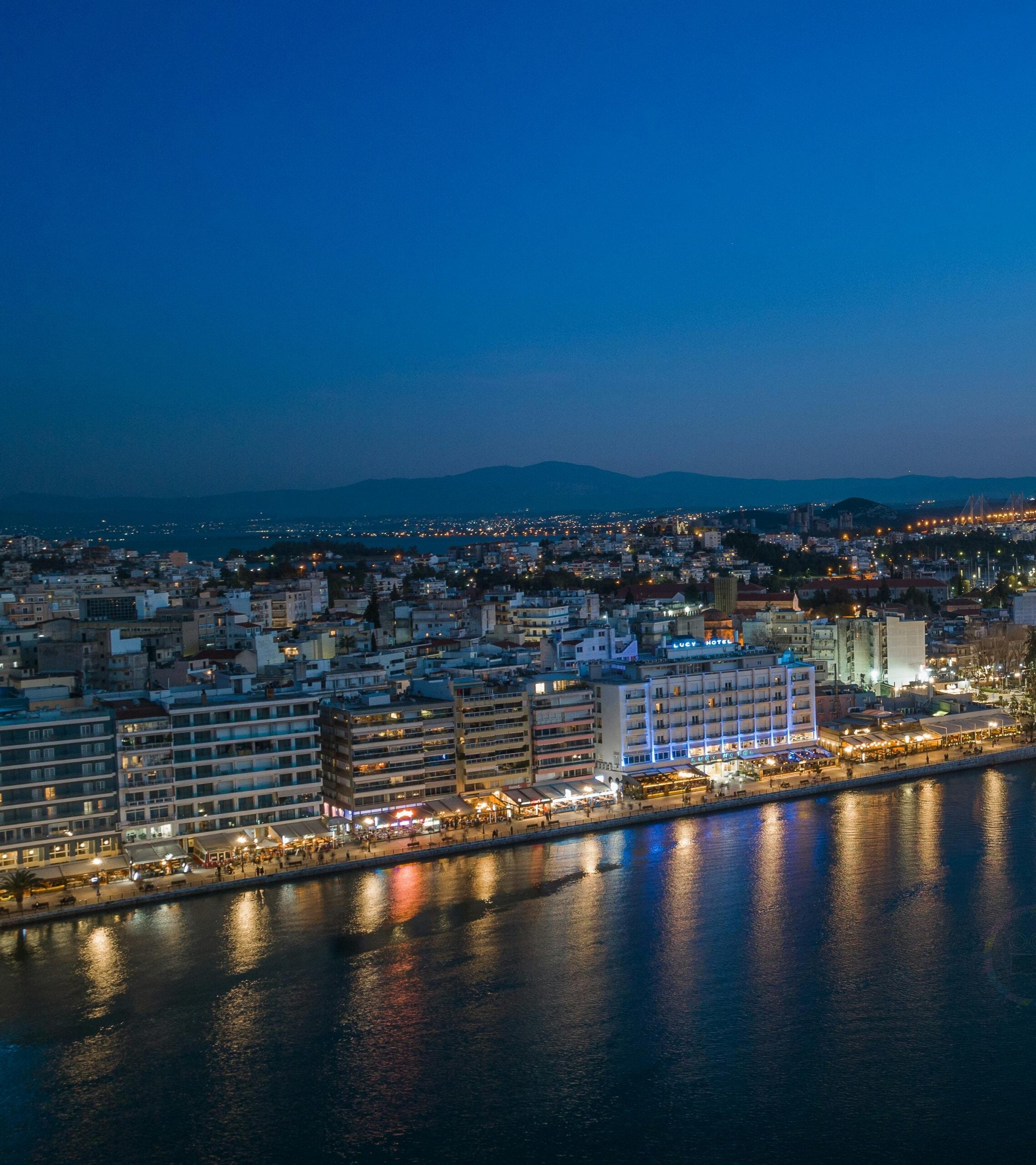 City waterfront at night with illuminated buildings reflecting on water and distant mountains in the background.