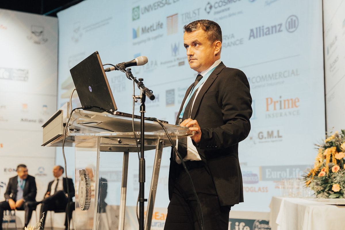 Speaker Presenting At A Conference With A Laptop And Microphone, Featuring Company Logos In The Background.
