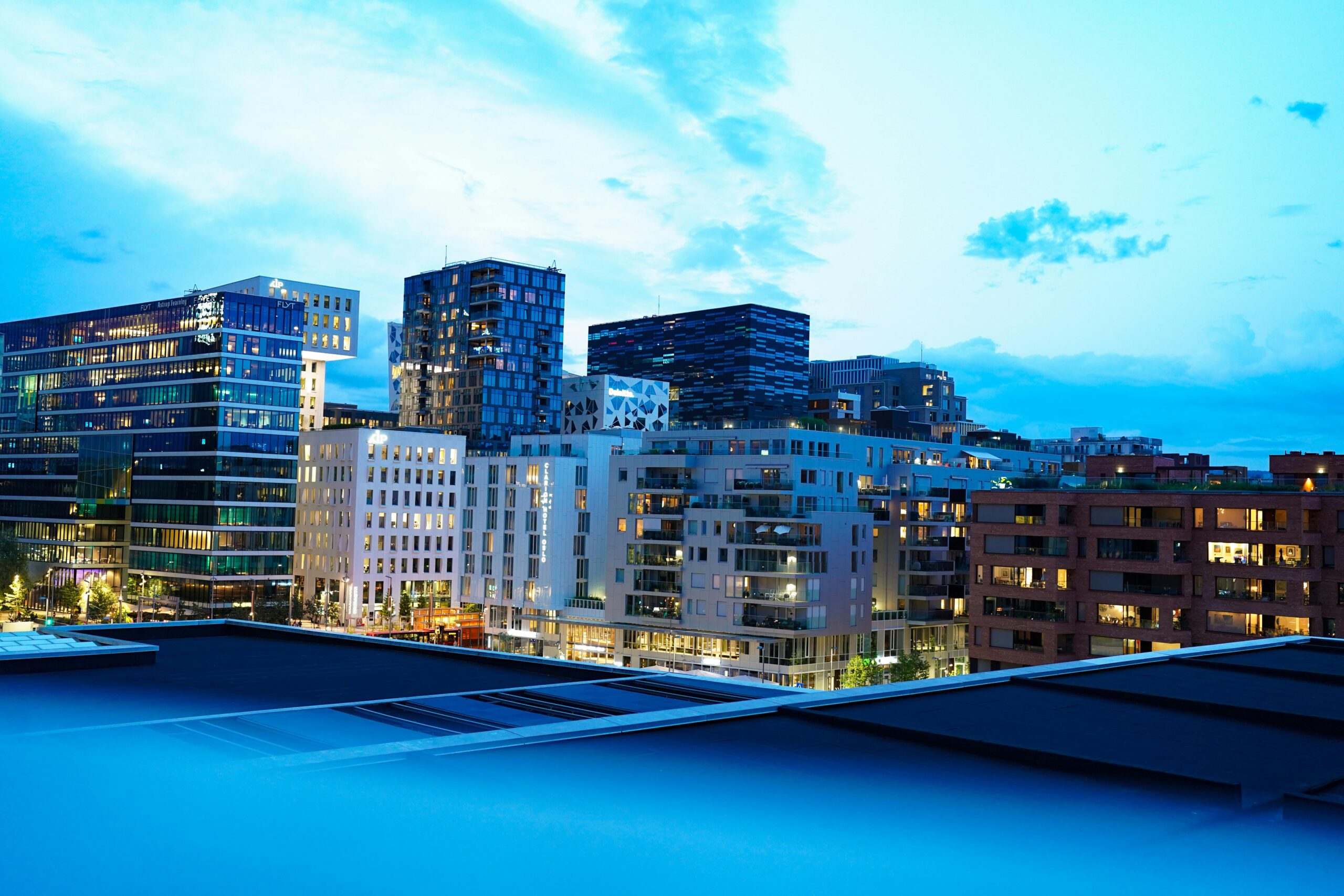 Urban skyline at dusk featuring modern buildings with illuminated windows and blue skies.