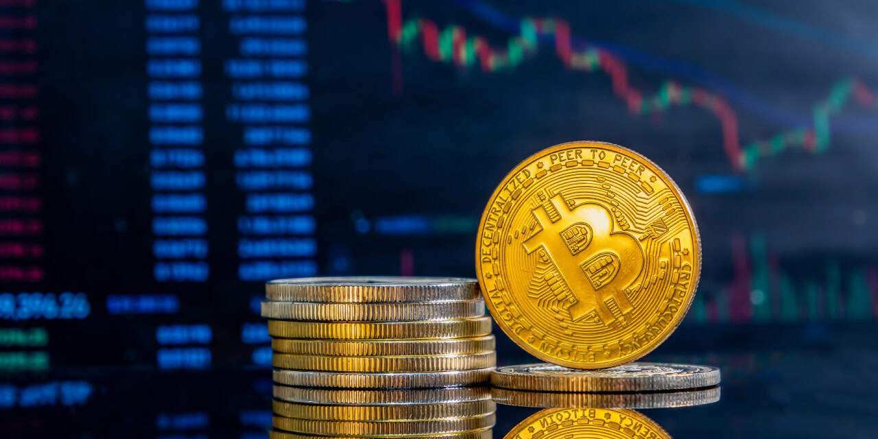 Stacks of golden Bitcoin coins are in the foreground, with a digital stock market chart in the blurry background, conveying a sense of financial trading.