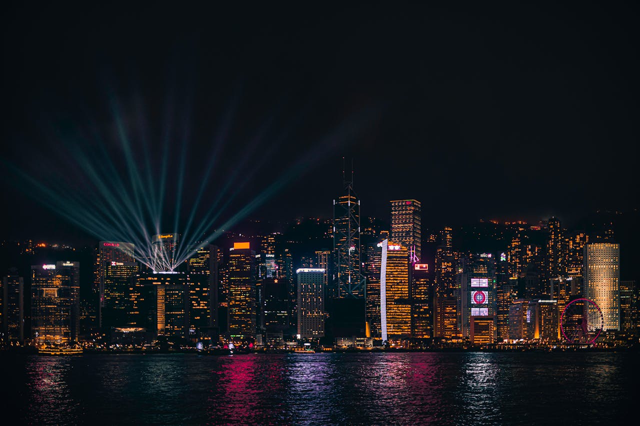 City skyline at night with illuminated skyscrapers and vibrant light beams reflecting on the water surface.