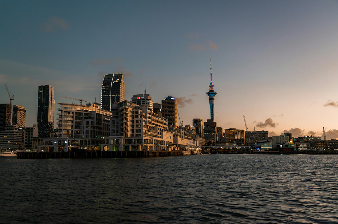 City skyline at sunset featuring modern buildings and the Sky Tower in Auckland, New Zealand.