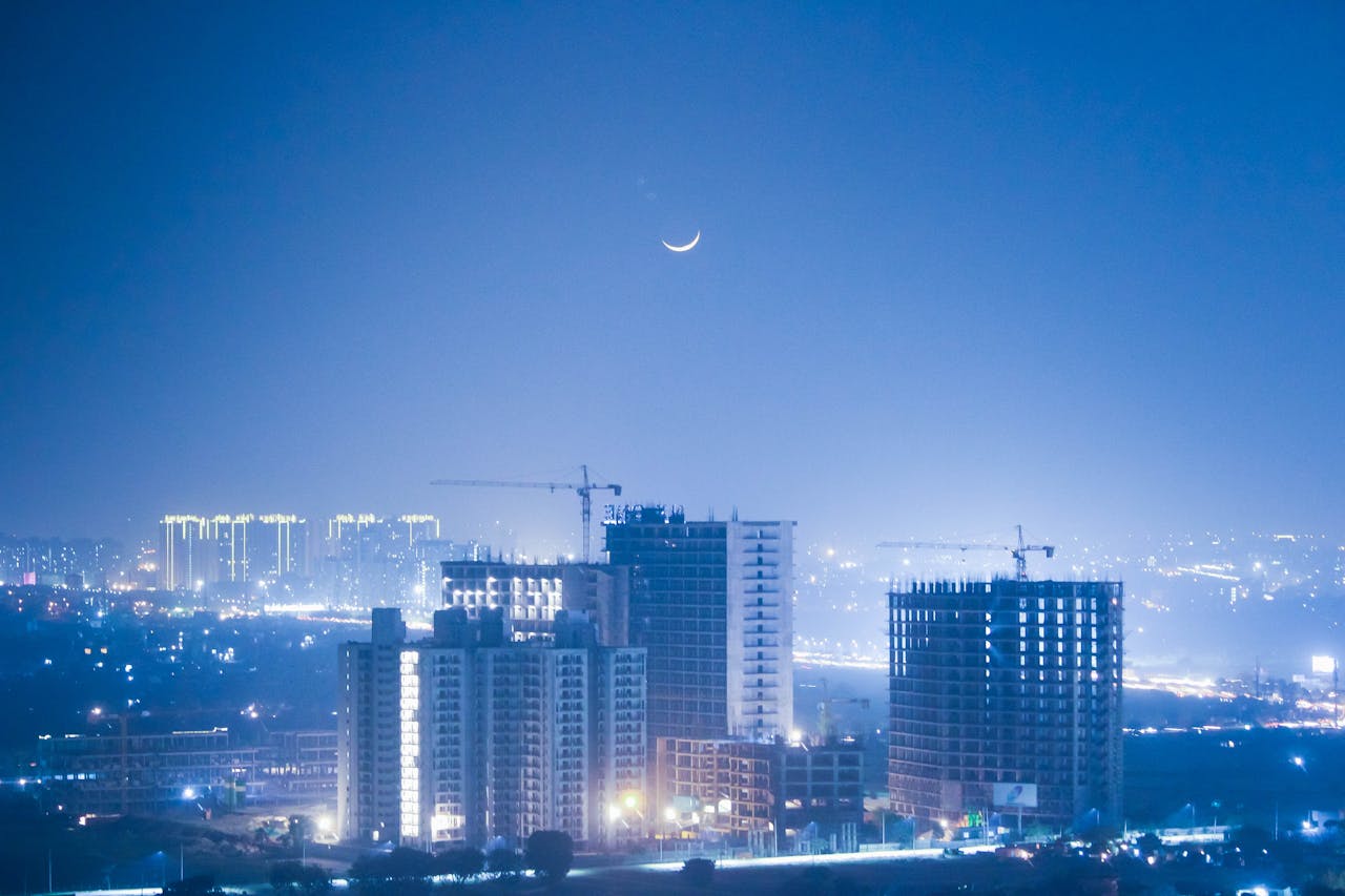 City skyline at night with construction sites and illuminated buildings under a crescent moon.