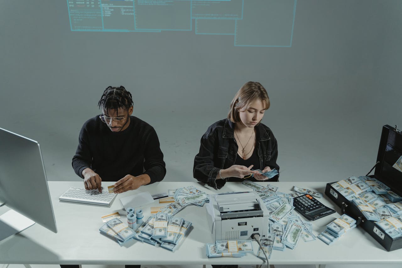 Man counting cash at desk while woman organizes money, with a printer and calculator on the table.