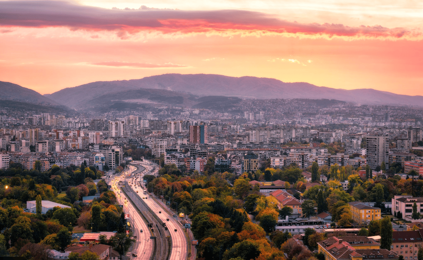 Cityscape view during sunset with skyline, mountains, and urban road featuring vehicles and tree-lined areas.