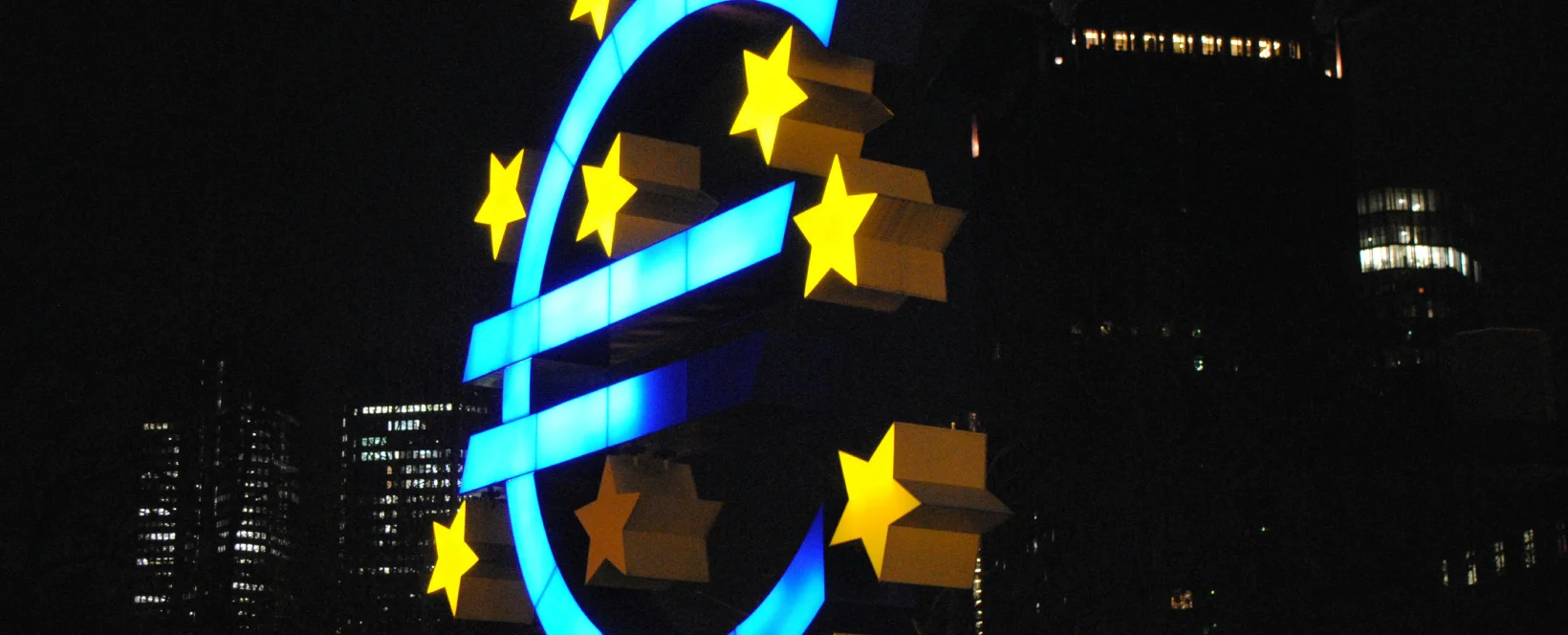 Euro symbol illuminated at night with stars, surrounded by city buildings in a legal or financial district.