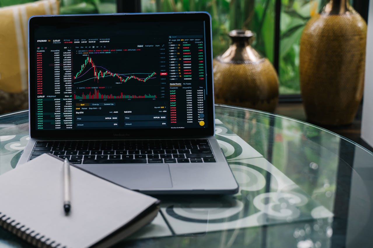 Laptop displaying financial market data and graphs on a glass table beside a notebook and pen.