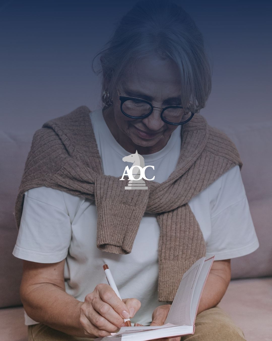 Senior individual taking notes while reviewing a booklet, seated comfortably on a couch with a sweater draped over shoulders.