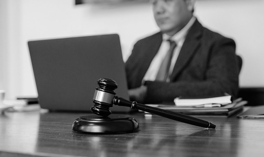 Lawyer seated at desk with laptop, gavel and pen on table, reviewing legal documents in office setting.