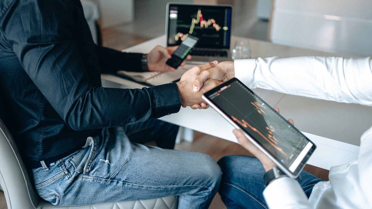 Business partners shaking hands during a meeting, with digital devices displaying financial data on the table.