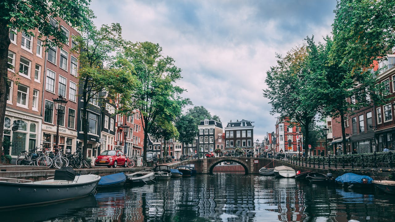 Canal lined with historic buildings, boats, and trees under a cloudy sky in an urban setting.