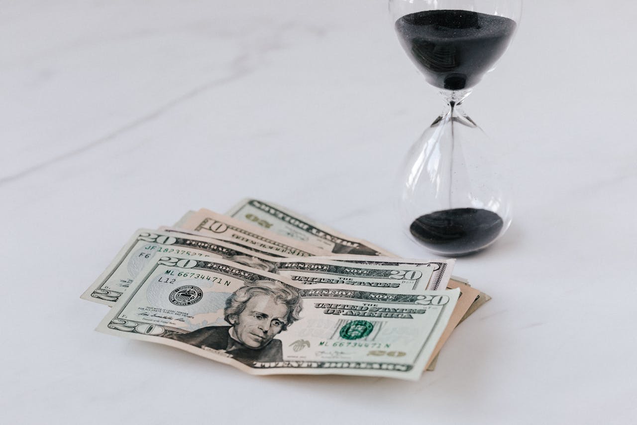 Stack of US dollar bills with a black sand hourglass on a white surface.