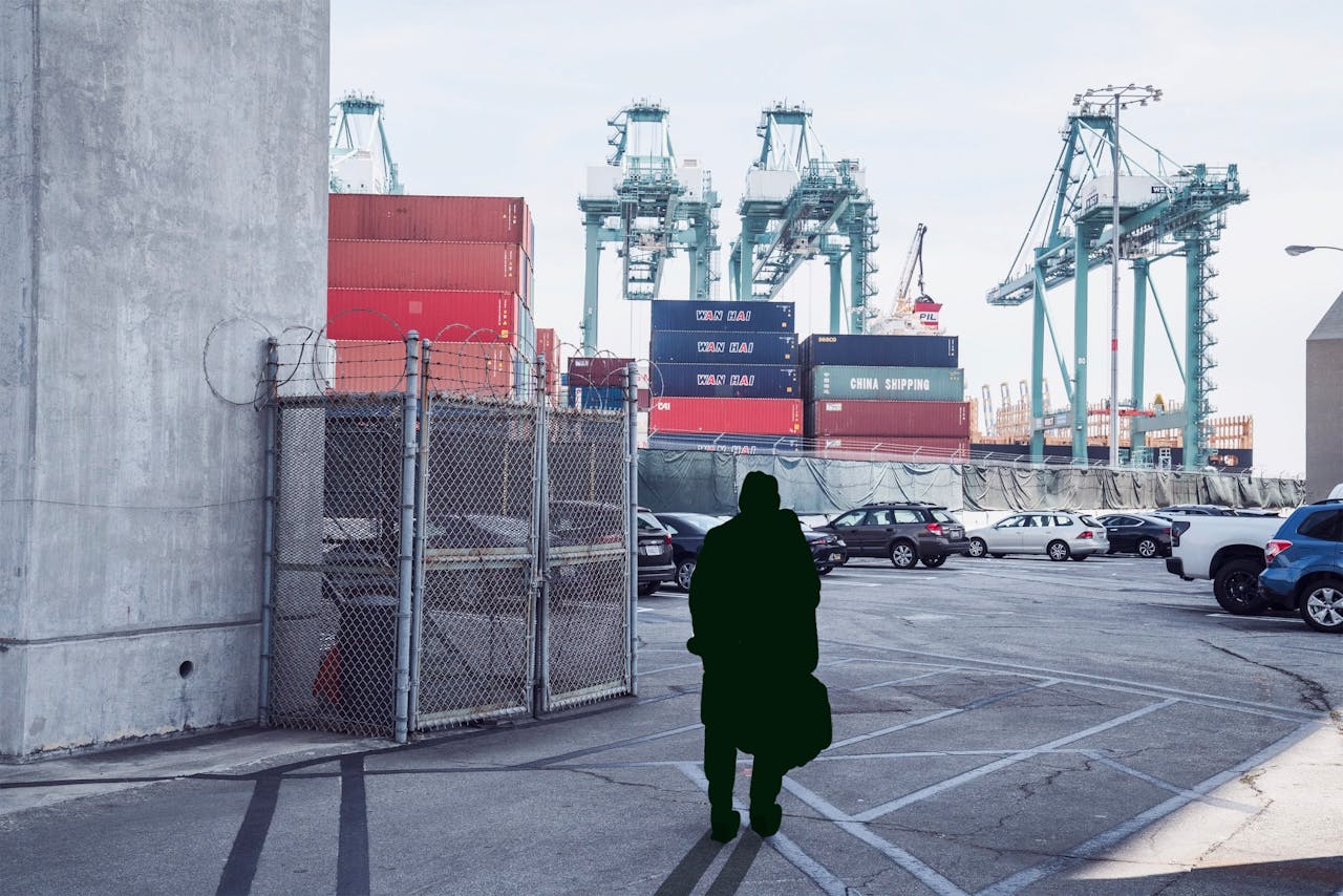 Person walking in a shipping yard, with containers and cranes in the background.