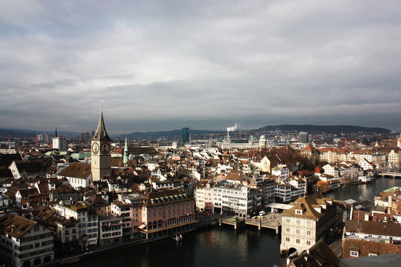 Cityscape of Zurich featuring historic buildings, a river, and a clock tower against a cloudy sky.