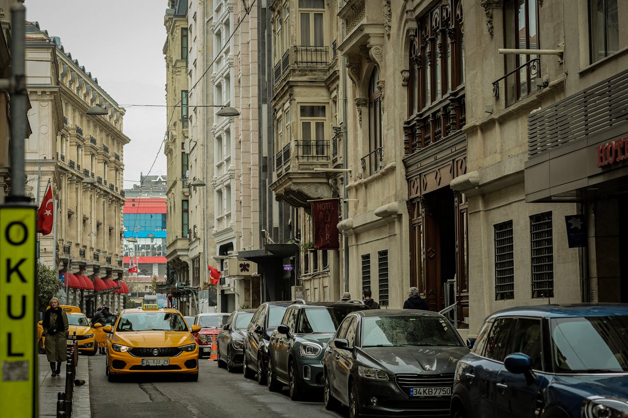 Narrow city street lined with parked cars and a yellow taxi, featuring historic buildings and Turkish flags.
