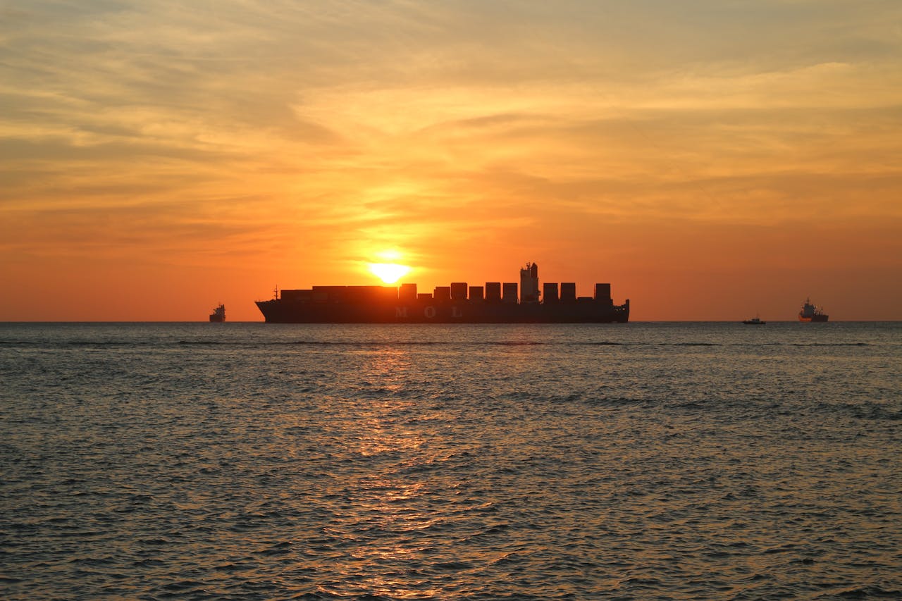 Container ship silhouetted against a sunset on the ocean, with smaller boats visible in the distance.