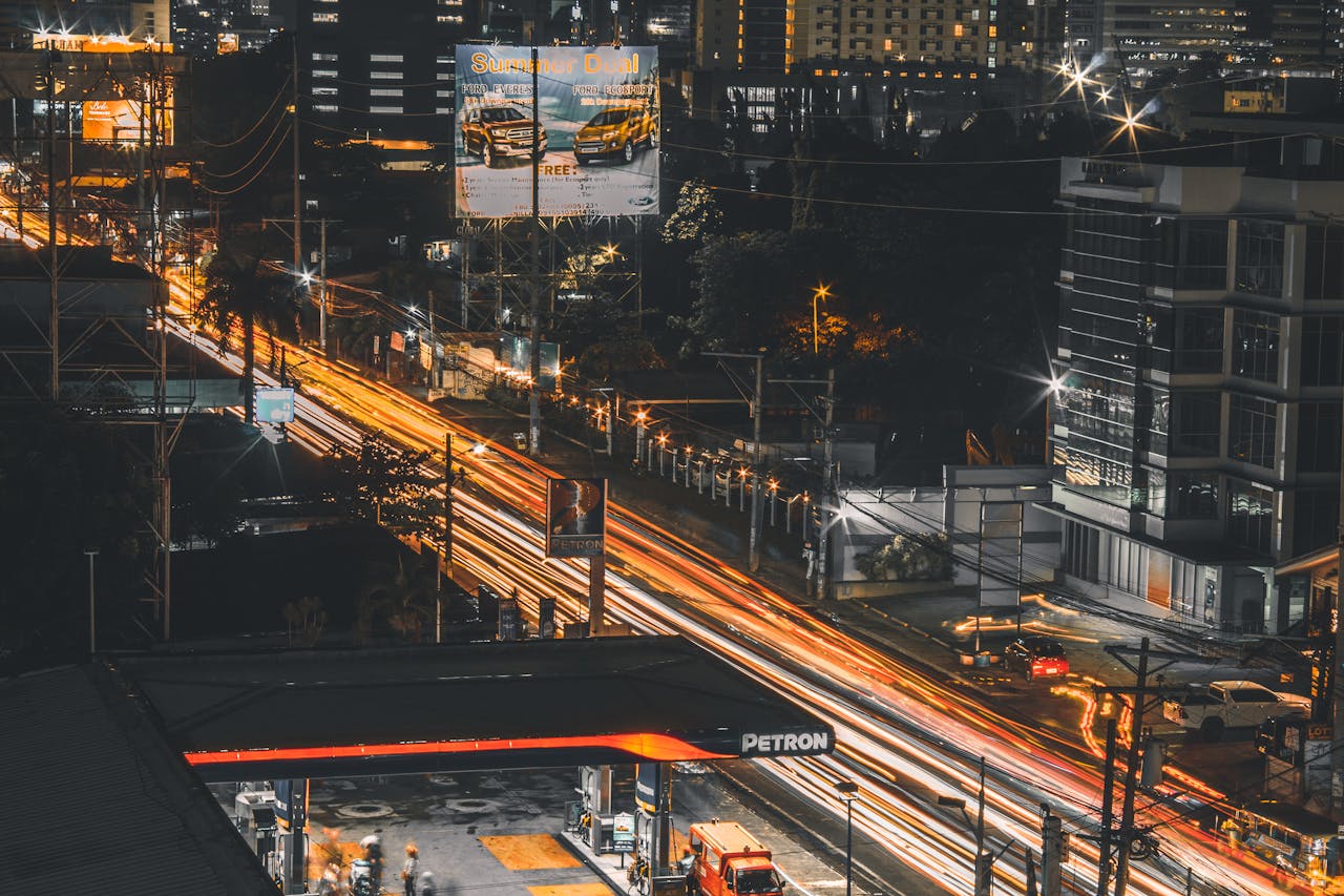 Urban street scene at night with light trails from vehicles moving past a gas station and modern buildings in the background.