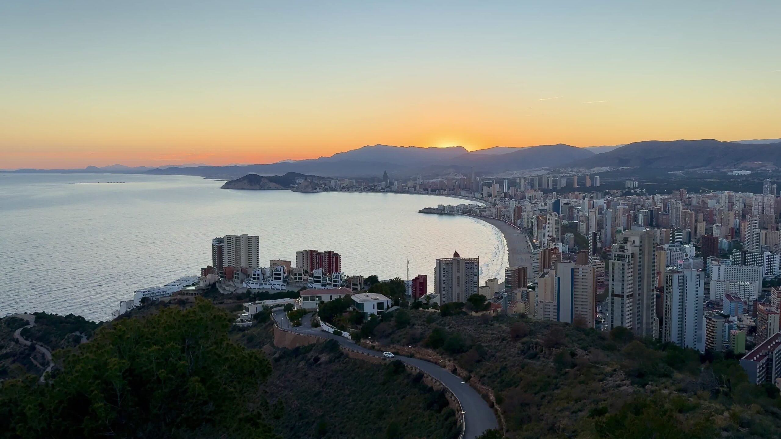 Coastal city skyline with high-rise buildings along the shore during sunset. Mountains in the background.
