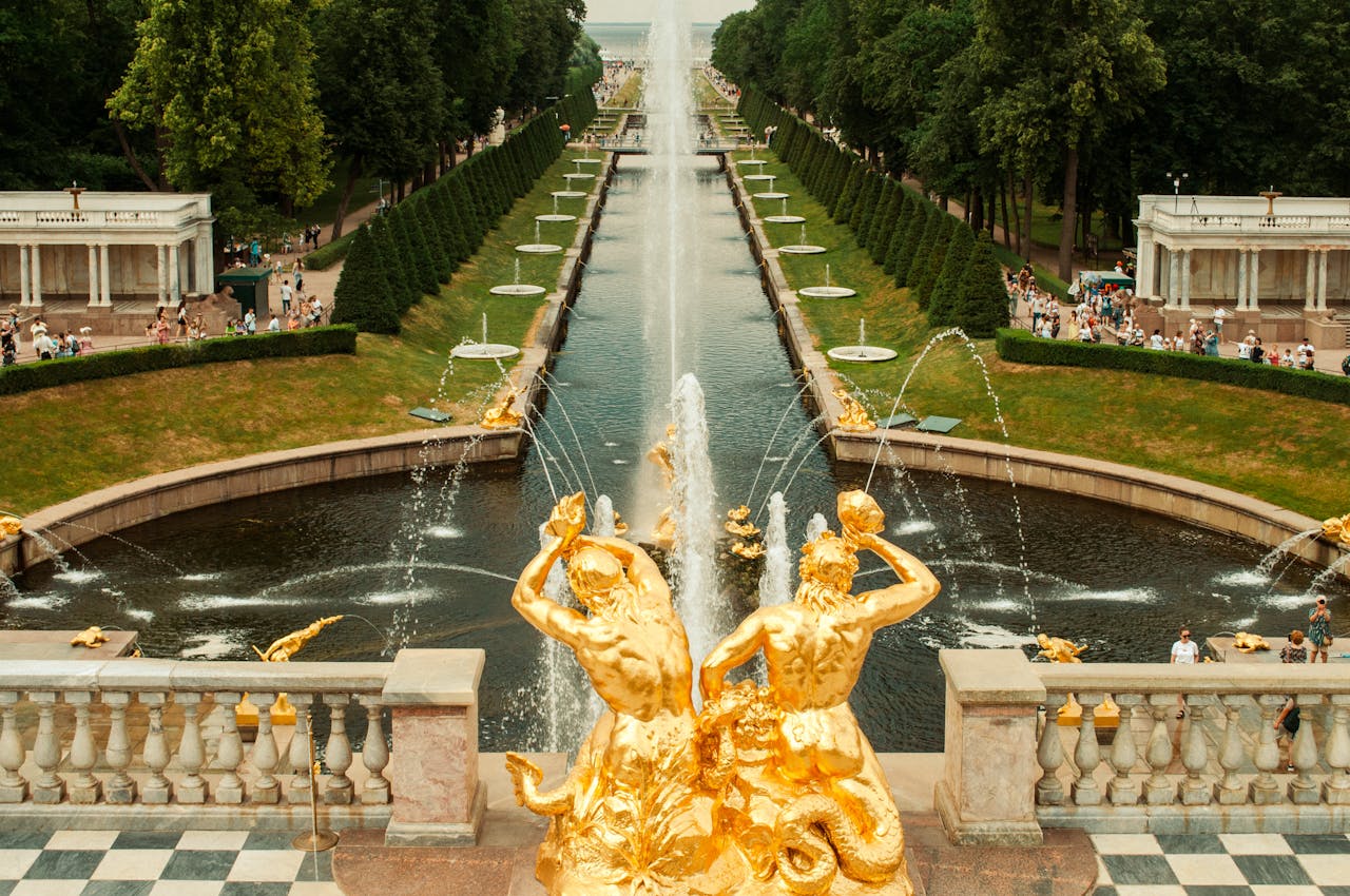 Golden statues with water fountains in a landscaped park filled with visitors enjoying the scenery.