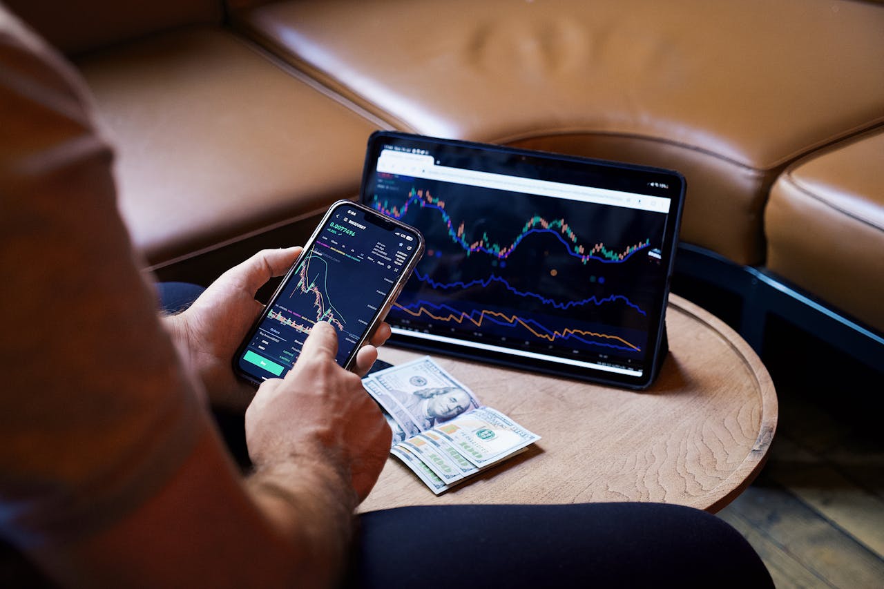 Person analyzing financial data on a smartphone and laptop, with cash on a wooden table.